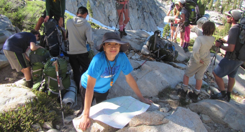 A young person leans against a rock on which a map is resting and smiles at the camera. Other outward bound students and their packs are pictured in the background. 